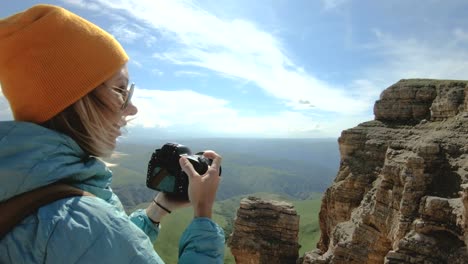 Blonde-girl-photographer-in-the-cap-takes-a-photo-on-her-digital-camera-with-a-background-of-rocks-in-the-Caucasus