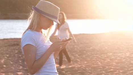 Schönes-Blondes-Mädchen-mit-Hut-Typen-eine-Nachricht-auf-ihr-Handy-am-Strand-bei-Sonnenuntergang.