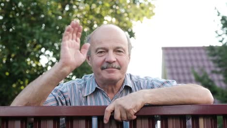 Friendly-caucasian-old-man-waving-hi-or-farewell,-isolated-outdoors-background-with-green-trees-and-fence