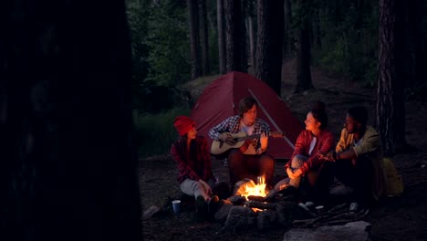 Multiracial-group-of-friends-hikers-is-playing-the-guitar,-singing-and-laughing-around-campfire-in-the-evening.-Summertime-fun,-nature-and-leisure-concept.