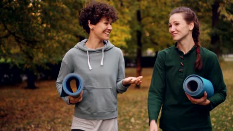 Schönes-Mädchen-geht-mit-Yoga-Lehrer-hält-Matten-reden-Gestikulieren-und-Lachen-im-Park-spazieren.-Lehrer-Schüler-Beziehung-und-Gespräch-Konzept.