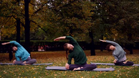 Instructora-de-yoga-de-mujer-es-mostrando-ejercicios-de-estiramiento-y-relajación-en-posición-de-loto-con-los-ojos-cerrados,-sentados-en-esteras-en-las-prácticas-al-aire-libre-en-el-parque.