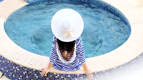 Mujer-de-elegancia-vista-trasera-de-alto-ángulo-con-sombrero-con-lazo-sentado-en-el-borde-de-la-piscina-o-jacuzzi