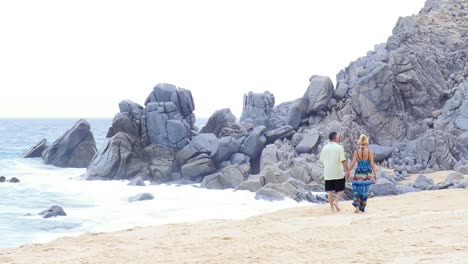 Eine-ältere-paar-Hände-halten,-zu-Fuß-am-Strand-entlang-in-vor-großen-Felsen