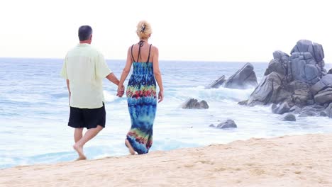 Eine-ältere-paar-Hände-halten,-zu-Fuß-am-Strand-entlang-in-vor-großen-Felsen