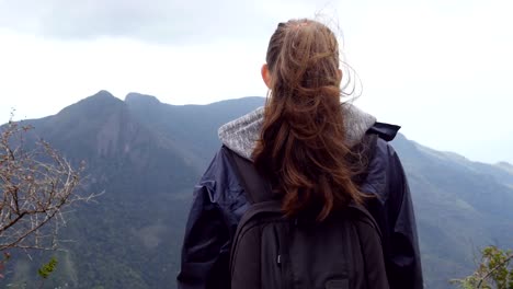 Unrecognizable-woman-tourist-standing-on-the-edge-of-beautiful-canyon-and-enjoying-landscapes.-Young-female-hiker-in-raincoat-with-backpack-reaching-up-top-of-mountain-and-raised-hands.-Rear-back-view