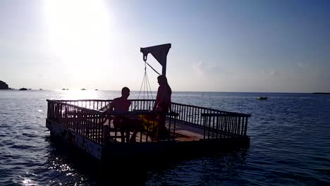 v03830-Aerial-flying-drone-view-of-Maldives-white-sandy-beach-2-people-young-couple-man-woman-relaxing-on-sunny-tropical-paradise-island-with-aqua-blue-sky-sea-water-ocean-4k-floating-pontoon-jetty-sunbathing-together