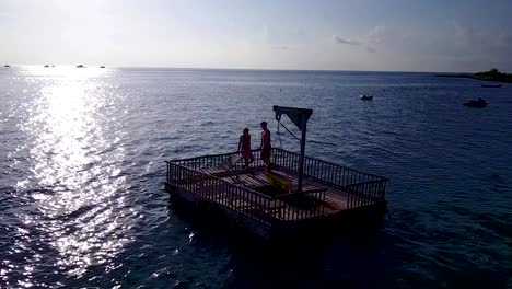 v03842-Aerial-flying-drone-view-of-Maldives-white-sandy-beach-2-people-young-couple-man-woman-relaxing-on-sunny-tropical-paradise-island-with-aqua-blue-sky-sea-water-ocean-4k-floating-pontoon-jetty-sunbathing-together