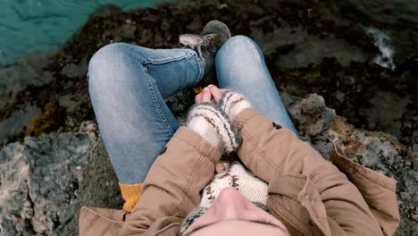 Close-up-view-of-young-woman-sitting-at-the-rock-near-blue-water.-Traveling-female-resting-after-hiking-alone