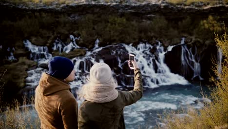 Young-traveling-couple-standing-in-mountains-valley-near-waterfall-and-talking-pictures-or-selfie-photos-on-smartphone