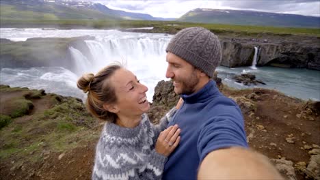 Travel-couple-fun-taking-selfie-photo-by-Godafoss-waterfall-on-Iceland-using-smartphone.-People-visiting-famous-tourist-attractions-and-landmarks-on-Route-1