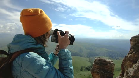 Blonde-girl-photographer-in-the-cap-takes-a-photo-on-her-digital-camera-with-a-background-of-rocks-in-the-Caucasus