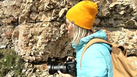 Blonde-girl-photographer-in-the-cap-takes-a-photo-on-her-digital-camera-with-a-background-of-rocks-in-the-Caucasus