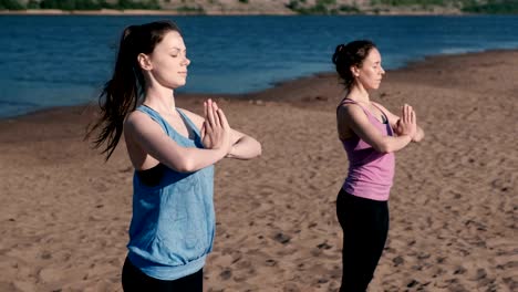 Two-woman-stretching-yoga-standing-on-the-beach-by-the-river-in-the-city.-Beautiful-city-view.-Namaste-pose.