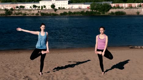 Two-woman-stretching-yoga-standing-on-the-beach-by-the-river-in-the-city.-Beautiful-city-view.-Balance-pose.