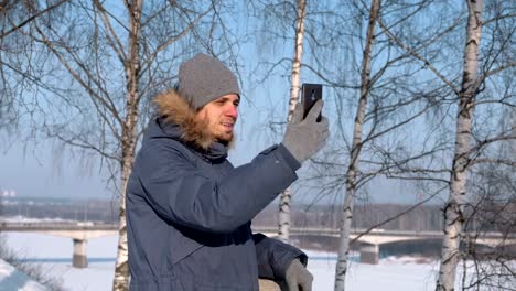 Mann-in-blau-Daunenjacke-mit-Fell-Kapuze-mit-Handy-für-Video-Chat-und-lächelnd-in-Winter-Park.