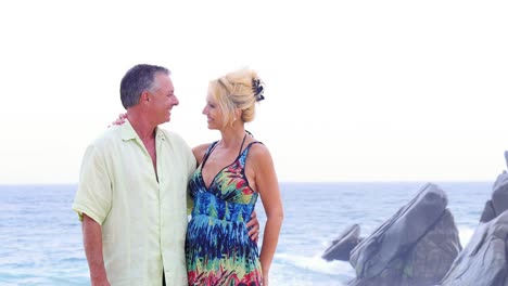 An-older-couple-smile-and-kiss-on-the-beach-with-waves-and-rocks-behind-them