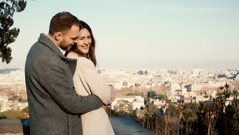 Romantic-date-of-young-couple.-Man-comes-to-woman-from-the-back-and-hugs-her.-Male-and-female-look-to-Rome,-Italy