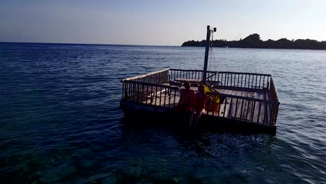 v03839-Aerial-flying-drone-view-of-Maldives-white-sandy-beach-2-people-young-couple-man-woman-relaxing-on-sunny-tropical-paradise-island-with-aqua-blue-sky-sea-water-ocean-4k-floating-pontoon-jetty-sunbathing-together