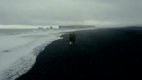 Aerial-view-of-the-hipster-couple-walking-on-the-shore-of-the-sea,-in-black-volcanic-beach-in-Iceland-together