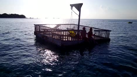 v03838-Aerial-flying-drone-view-of-Maldives-white-sandy-beach-2-people-young-couple-man-woman-relaxing-on-sunny-tropical-paradise-island-with-aqua-blue-sky-sea-water-ocean-4k-floating-pontoon-jetty-sunbathing-together