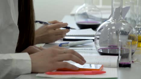 Los-estudiantes-en-el-trabajo-en-el-laboratorio-de-química-toman-notas-en-un-cuaderno.-Mujer-alumno-usando-el-teléfono-en-lección-de-ciencia.-Escritura-de-jóvenes-estudiantes-de-medicina