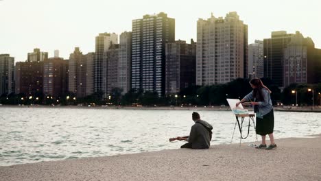 Hermosa-joven-pareja-en-la-orilla-del-lago-Michigan,-Chicago,-América.-Dibujo-de-mujer,-guitarra-juego-de-hombre