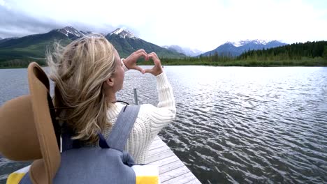 Young-woman-stands-on-wooden-jetty-by-the-lake,-she-makes.-heart-shape-frame