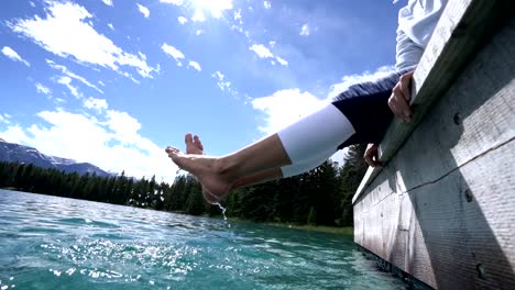 Woman's-feet-dangle-from-wooden-pier,-above-lake