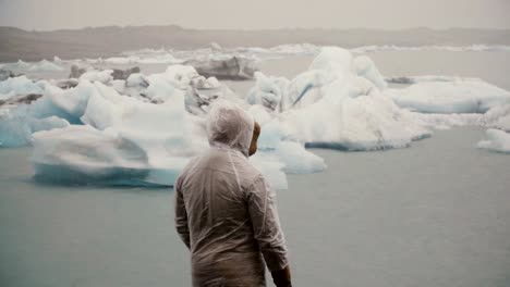Back-view-of-young-man-in-raincoat-standing-in-ice-lagoon-in-Iceland.-Tourist-exploring-the-famous-sight-alone