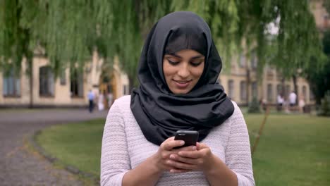 Young-beautiful-muslim-girl-in-hijab-is-typing-message-on-her-smartphone,-watching-at-camera-in-blue-weather,-communication-concept,-religious-concept