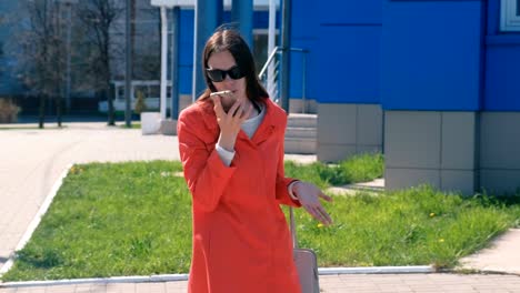 Young-brunette-woman-in-sunglasses-speaks-shouting-and-swearing-on-the-phone-beside-blue-building.