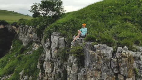 Young-hipster-woman-is-sitting-on-the-edge-on-the-grass-of-tall-wall-of-rock.-Aerial-view.-Drone-is-flying-slow-backward-from-model.-Establishing-revealing-shot