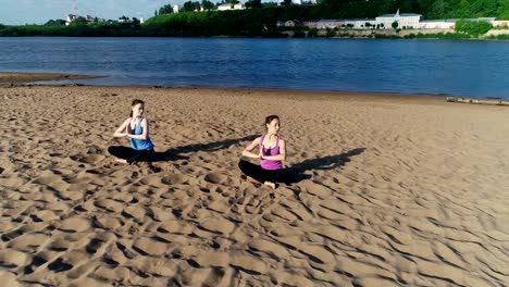 Two-women-stretching-yoga-on-the-sandy-beach-of-the-river.-Beautiful-city-view.