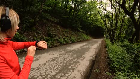 Young-attractive-woman-in-headphones-changing-the-settings-on-a-smart-watch-in-front-of-or-psole-jogging-on-the-road-in-a-green-forest