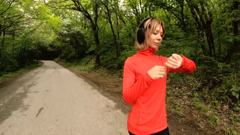 Young-attractive-woman-in-headphones-changing-the-settings-on-a-smart-watch-in-front-of-or-psole-jogging-on-the-road-in-a-green-forest