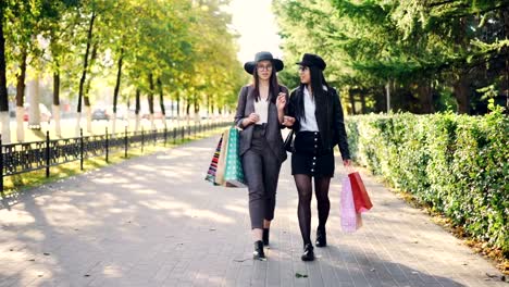 Dolly-shot-of-happy-girls-shopaholics-walking-in-the-street-carrying-shopping-paper-bags-and-talking-discussing-new-clothes-collection.-Youth,-leisure-and-consumerism-concept.