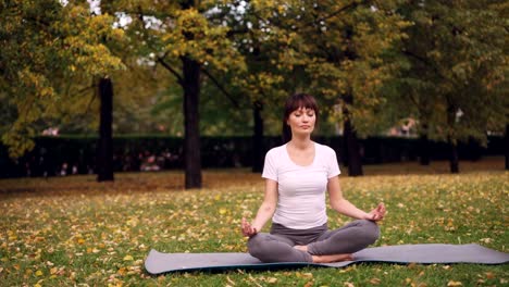 Attractive-young-lady-is-sitting-in-lotus-position-on-yoga-mat-in-park-holding-hands-in-mudra-on-knees-and-breathing-relaxing-after-practice.-Meditation-and-nature-concept.