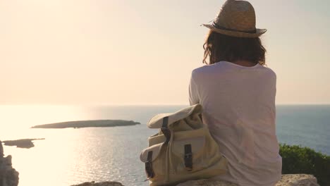 Portrait-of-a-beautiful-young-tourist-(girl),-in-a-white-dress,-looks,-with-a-backpack,-in-a-straw-hat,-walking-along-rocks,-sea-background-and-mountains.