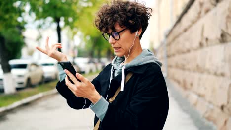 Young-woman-with-curly-hair-and-glasses-is-talking-to-friends-online-standing-outdoors-and-using-smartphone-and-earphones.-Gadgets,-internet-and-communication-concept.