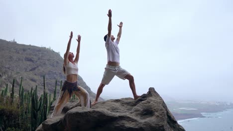 Un-hombre-y-una-mujer-de-pie-en-el-borde-de-un-acantilado-con-vistas-al-océano-levantan-sus-manos-y-aspirar-el-aire-del-mar-durante-yoga