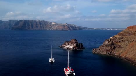 Flight-over-small-island-church-(Nisis-Agios-Nikolaos)-near-Oia-town,-Santorini-island,-Greece