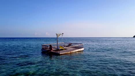 v03834-Aerial-flying-drone-view-of-Maldives-white-sandy-beach-2-people-young-couple-man-woman-relaxing-on-sunny-tropical-paradise-island-with-aqua-blue-sky-sea-water-ocean-4k-floating-pontoon-jetty-sunbathing-together