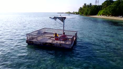 v03843-Aerial-flying-drone-view-of-Maldives-white-sandy-beach-2-people-young-couple-man-woman-relaxing-on-sunny-tropical-paradise-island-with-aqua-blue-sky-sea-water-ocean-4k-floating-pontoon-jetty-sunbathing-together