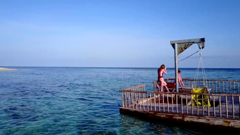 v03833-Aerial-flying-drone-view-of-Maldives-white-sandy-beach-2-people-young-couple-man-woman-relaxing-on-sunny-tropical-paradise-island-with-aqua-blue-sky-sea-water-ocean-4k-floating-pontoon-jetty-sunbathing-together