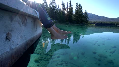 Woman's-feet-dangle-from-wooden-pier,-above-lake