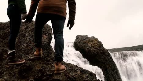 Young-man-and-woman-standing-on-a-hill-after-hiking-and-looking-on-a-powerful-waterfall-in-Iceland,-famous-sight