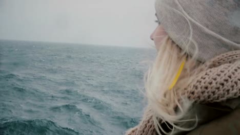 Close-up-view-of-young-blonde-woman-traveling-on-ship.-Attractive-female-looking-on-sea-and-waves,-hair-waving-on-wind