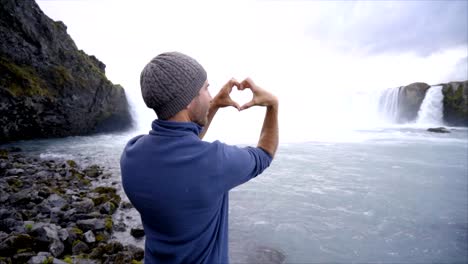 Junger-Mann-in-Island-machen-Herz-Formrahmen-Finger-auf-spektakulären-Wasserfall.-In-Island,-Frühling-gedreht.-Menschen-Reisen-Liebe-Lifestyle-Konzept