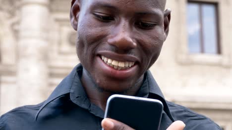 Sonriente-hombre-joven-africano-americano-tecleando-en-su-teléfono-inteligente-al-aire-libre